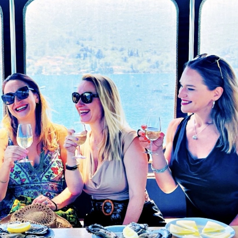 Three women toasting with white wine over oysters and cheese on a boat by the sea