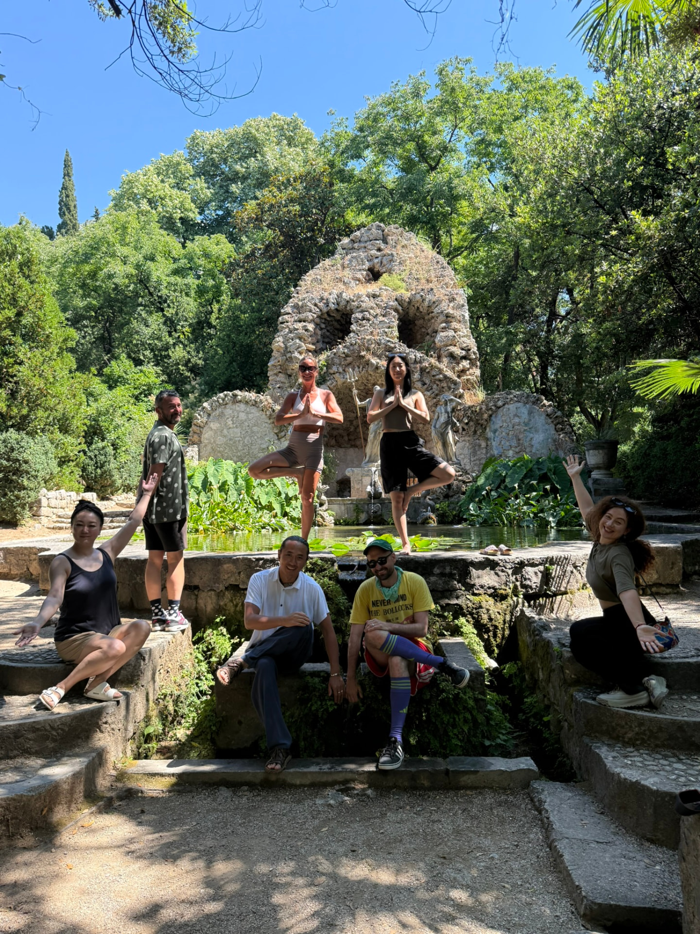 Guests gathered in the sunlight at the Neptune Fountain grotto in Trsteno Arboretum, two of them holding yoga tree poses by the pond